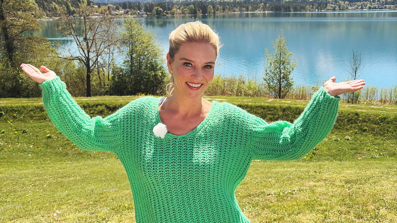 Christine Reiler steht auf einer Wiese am Faaker See und hebt lächelnd ihre Hände.