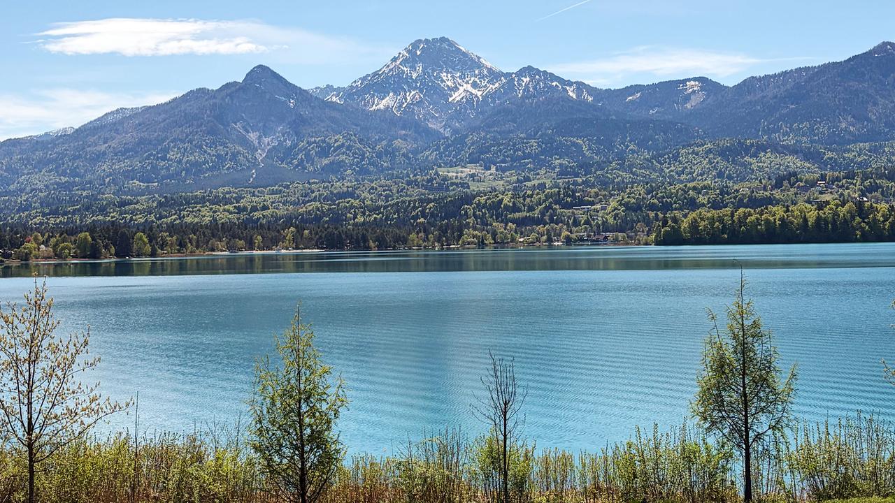 Faaker See mit Berglandschaft und bewaldeten Hängen und schneebedeckten Gipfeln im Hintergrund