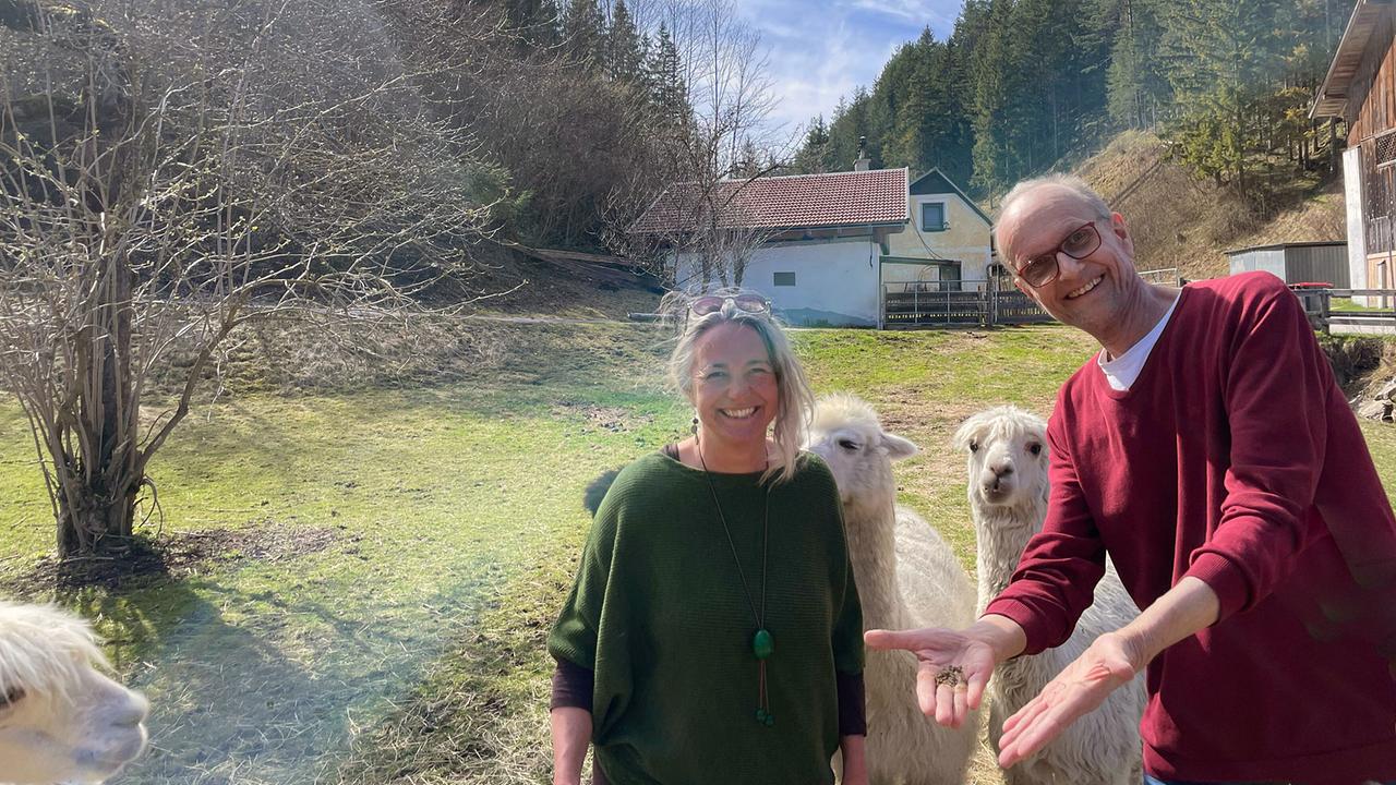 Barbara Enk vom Auszeithof Enk steht mit dem Besucher Thomas Heiland auf einer Wiese neben neugierigen Lamas oder Alpakas in alpiner Umgebung.