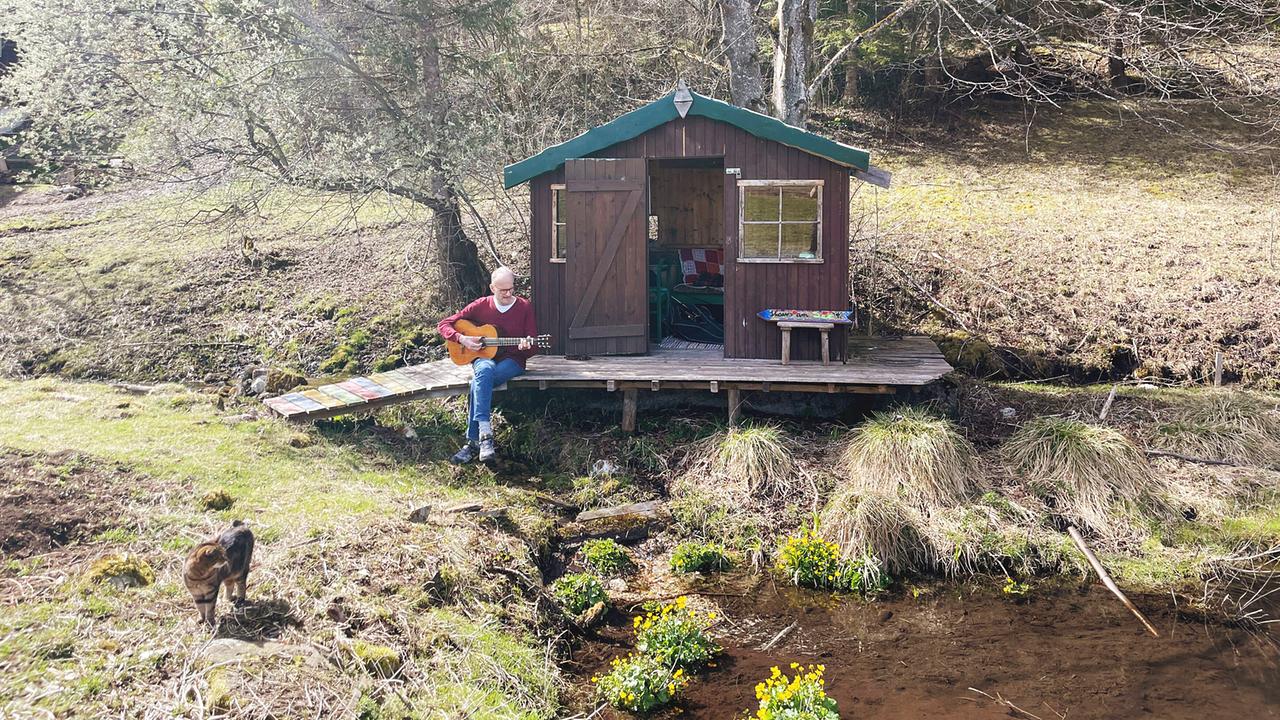 Krebspatient Thomas Heiland  am Auszeithof Enk  sitzt vor einer kleinen Holzhütte in der Natur und spielt Gitarre, während ein Tier im Vordergrund steht.