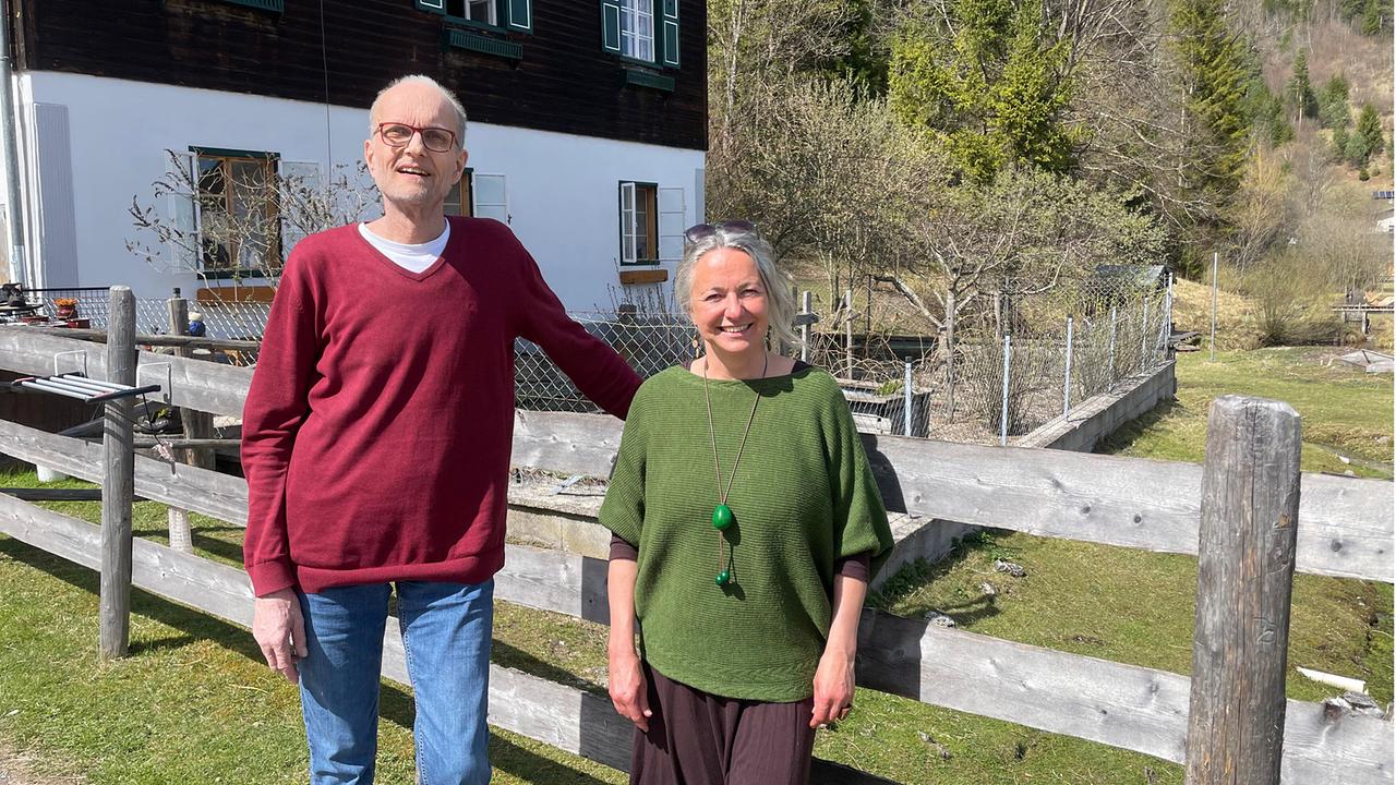 Barbara Enk und Besucher Thomas Heiland stehen am Auszeithof Enk vor einem ländlichen Haus hinter einem Holzzaun in sonniger Umgebung.