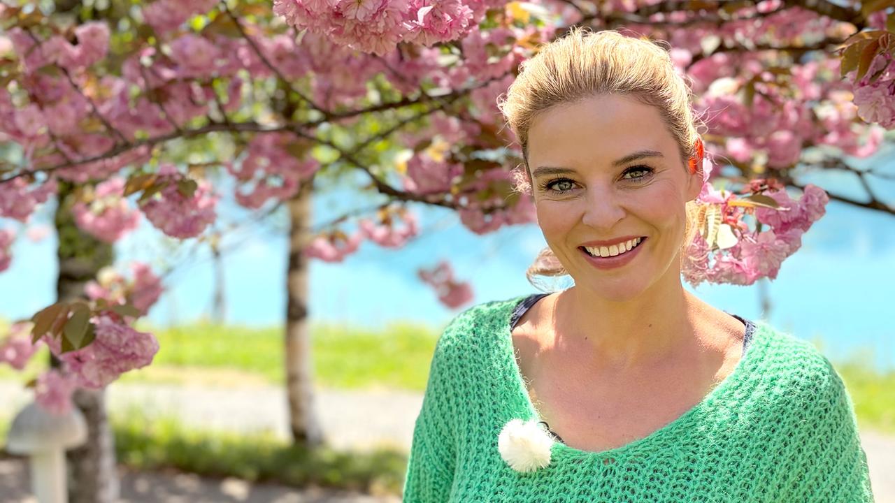 Christine Reiler steht im Frühling unter rosa blühenden Bäumen an einem sonnigen Weg am Faaker See.