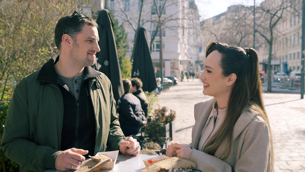 © ORF/TVfriends Ferdinand Seebacher und Sasa Schwarzjirg stehen an einem Stehtisch im Freien, essen belegte Brote und unterhalten sich in einer städtischen Umgebung.