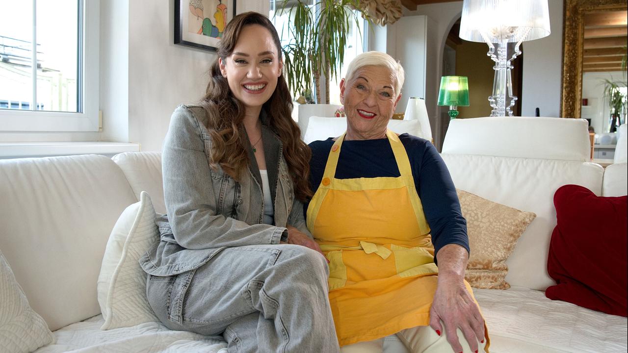 Sasa Schwarzjirg sitzt neben Jazz Gitti, die eine gelbe Kochschürze trägt, auf einem weißen Sofa in einem gemütlich eingerichteten Wohnzimmer.