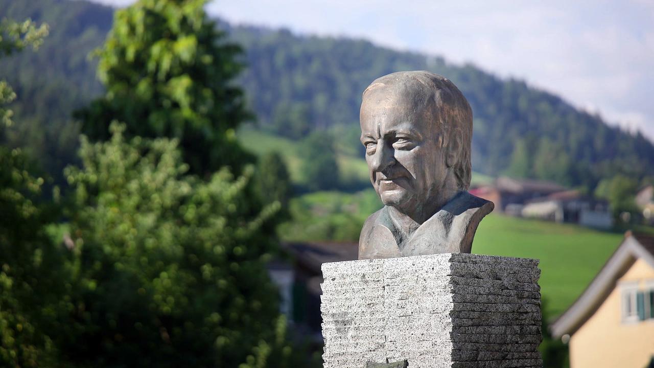 © ORF Eine Büste von Hermann Gmeiner steht auf einem Sockel vor einer hügeligen Landschaft.