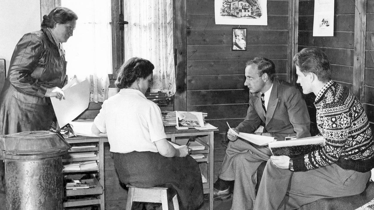 © ORF/SOS Kinderdorf Schwarz-Weiß-Foto von Hermann Gmeiner, der mit drei weiteren Menschen in einem urigen Innenraum in Innsbruck 1949 sitzt.