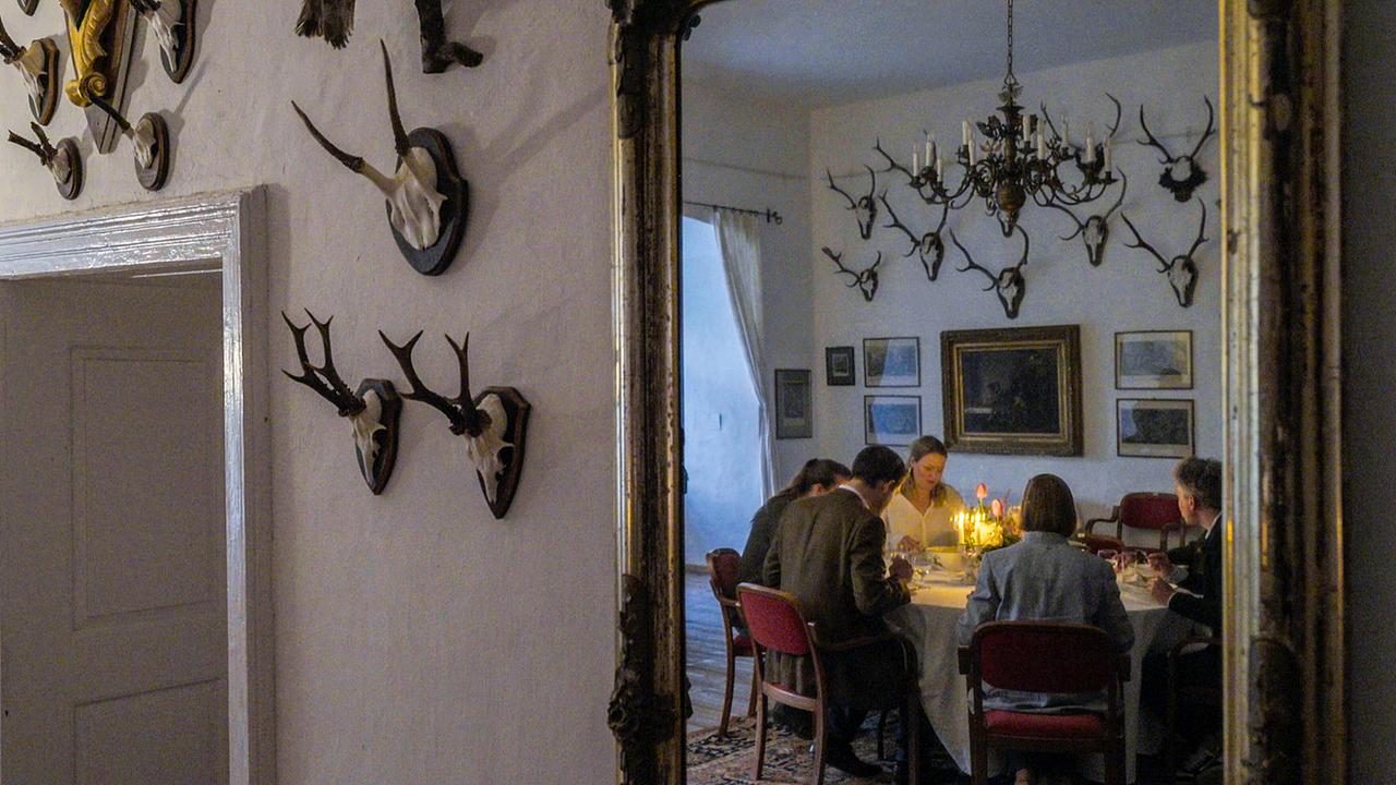 © ORF/Interspot In einem mit Jagdtrophäen geschmückten Speisezimmer sitzt Johann-Philipp Spiegelfeld mit Anna Schlanitz-Bolldorf und ihrer Familie bei Kerzenlicht an einem gedeckten Tisch.