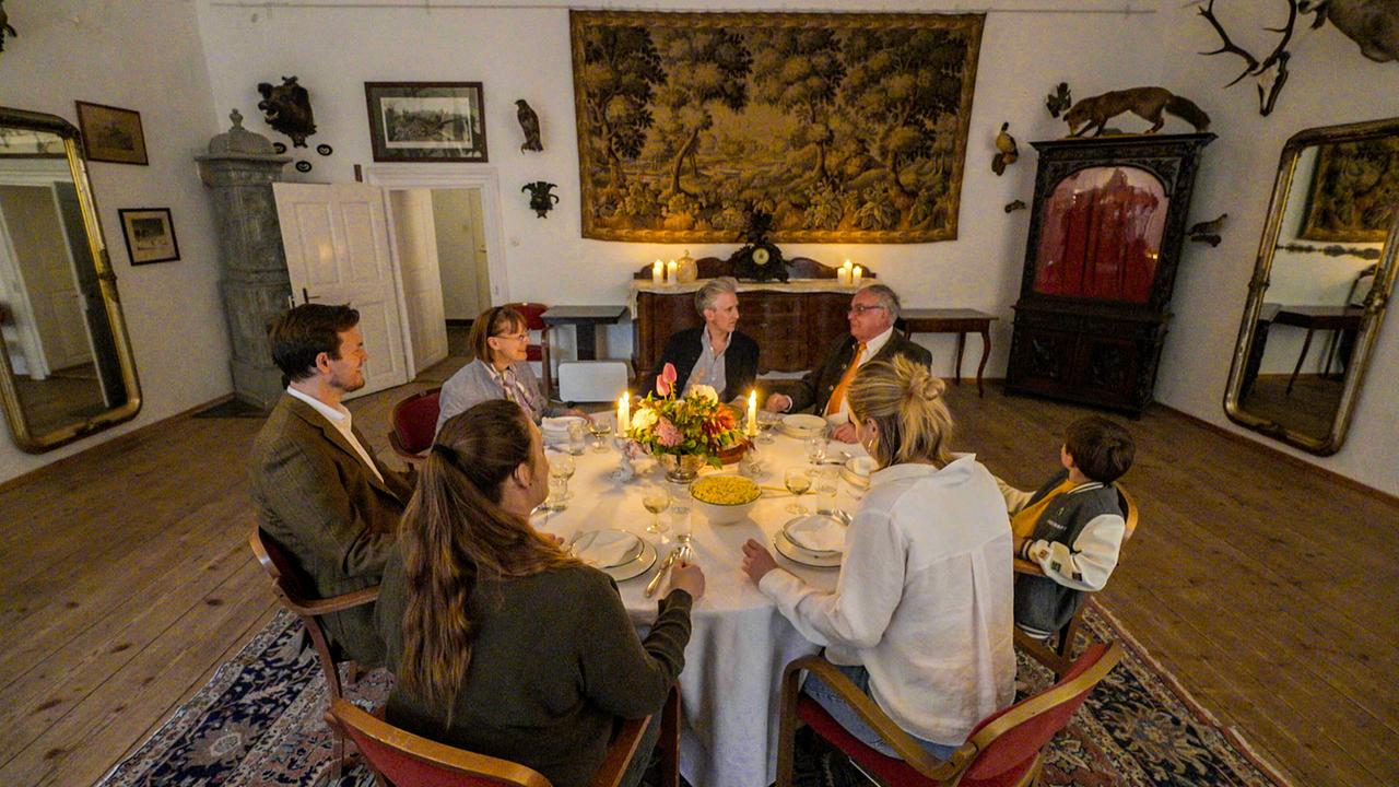 © ORF/Interspot Johann-Philipp Spiegelfeld speist mit Anna Schlanitz-Bolldorf und ihrer Familie bei Kerzenlicht in einem historischen Raum mit Wandteppich, antiken Möbeln und zahlreichen Jagdtrophäen.
