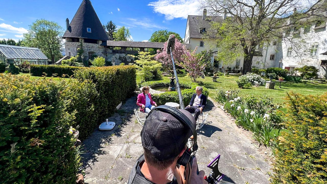 © ORF/Interspot/Philip Pürcher, Carlo Hofmann Ein Kameramann filmt ein Gespräch von Johann-Philipp Spiegelfeld und Anna Schlanitz-Bolldorf, die in Gartensesseln im sonnigen Schlossgarten von Kobersdorf sitzen.