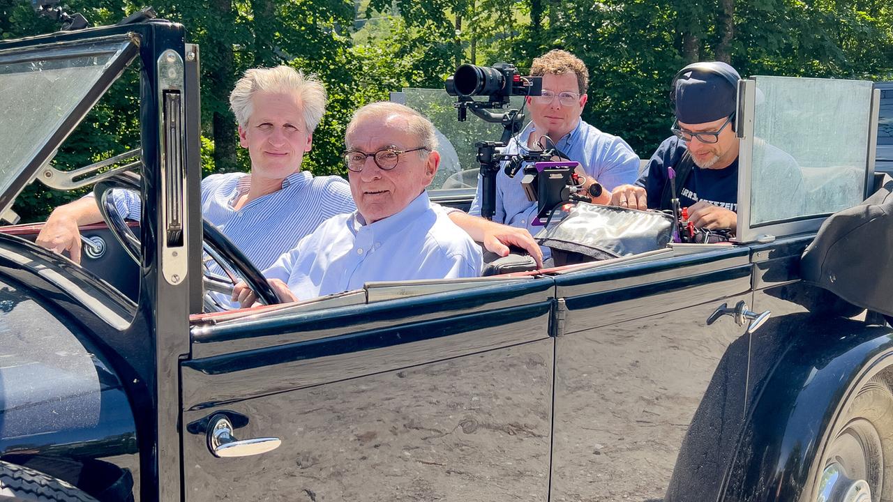 Johann-Philipp Spiegelfeld sitzt neben Wolfgang Boesch in einem offenen schwarzen Oldtimer mit der Aufschrift „Burg Strechau“, begleitet von Kameramann Carlo Hofmann  und Andreas Hagemann, zuständig für Ton, auf der Rückbank.