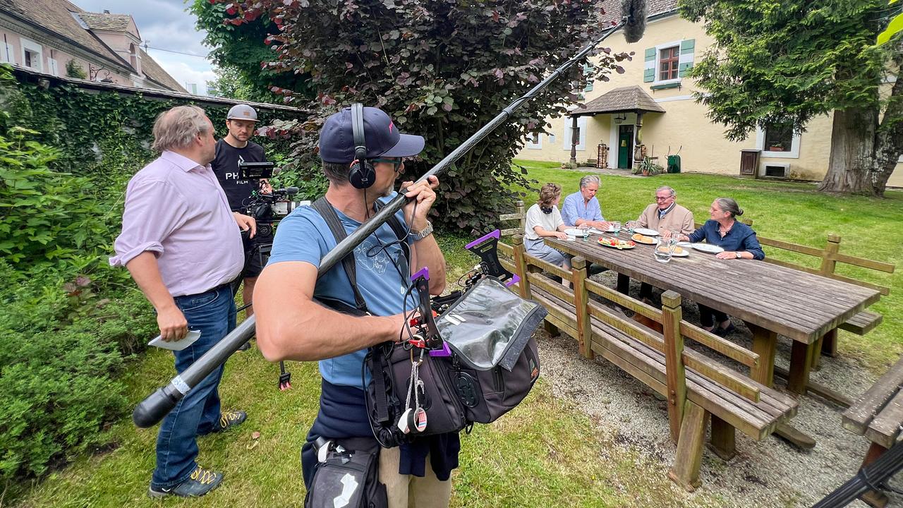 Bernadette Boesch, Johann-Philipp Spiegelfeld, Wolfgang Boesch und Marianne Boesch-Leibrecht sitzen beim Essen am Gartentisch  vor dem Burggebäude bei den Dreharbeiten mit dem Kamerateam mit Tonangel und Kamera im Vordergrund.