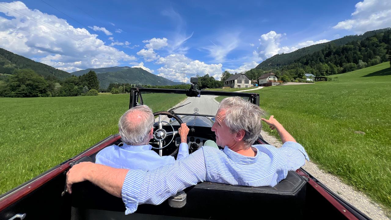 Johann-Philipp Spiegelfeld sitzt neben Wolfgang Boesch in einem offenen Oldtimer und sie fahren durch eine grüne Landschaft mit Bergen und blauem Himmel.