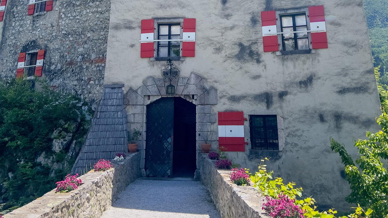 Burg Strechau mit dem Burgeingang mit weiß-grauer Fassade, roten Fensterläden und einem Turm mit Uhr über einer kleinen Brücke