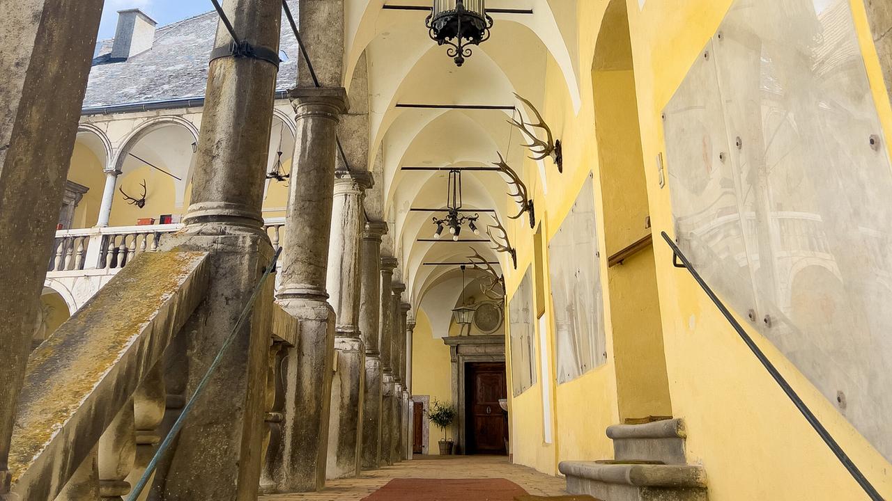Ein gelber Arkadengang in der Burg Strechau mit steinernen Säulen, Hängeleuchtern und an den Wänden befestigten Geweihen