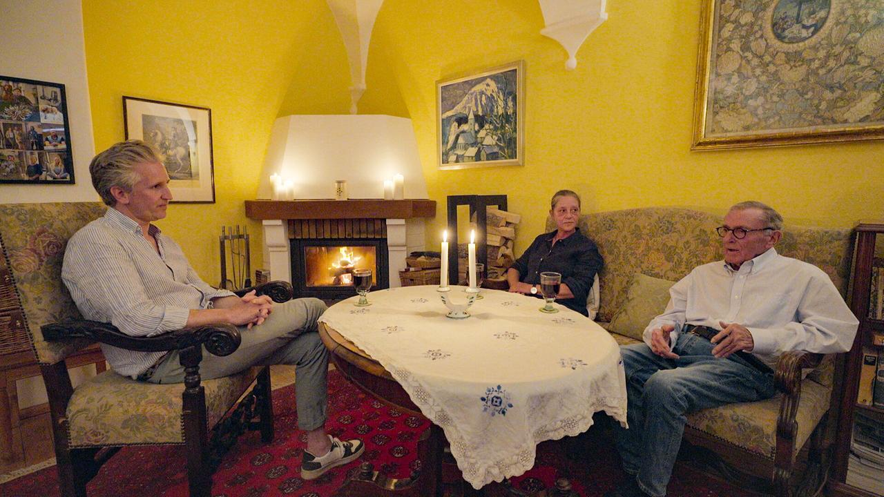 Johann-Philipp Spiegelfeld, Wolfgang Boesch und Marianne Boesch-Leibrecht sitzen in einem gemütlichen, gelb gestrichenen Zimmer bei Kerzenlicht und Kaminfeuer an einem Tisch.