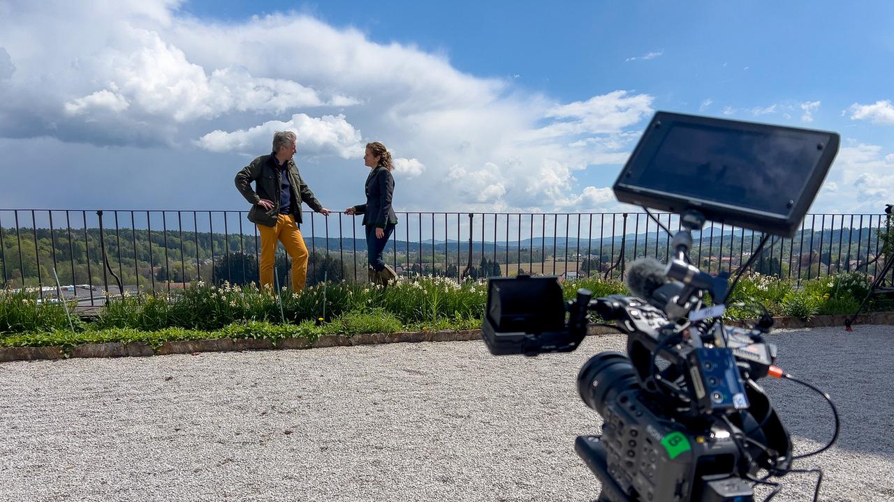 © ORF/Interspot/Philip Pürcher, Carlo Hofmann Johann-Philipp Spiegelfeld steht mit Schlossbesitzerin Angelika Meran bei einem Gespräch auf einer Aussichtsterrasse mit Kamera im Vordergrund.