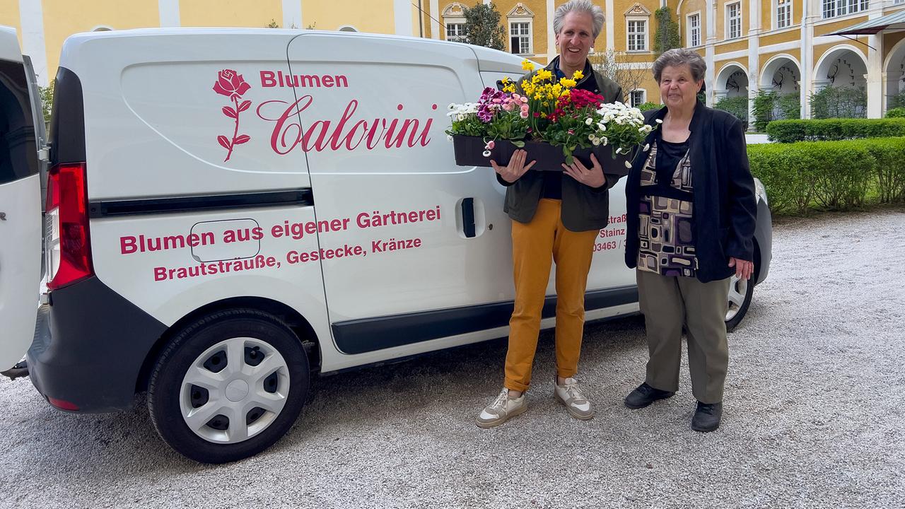 © ORF/Interspot/Philip Pürcher, Carlo Hofmann Johann-Philipp Spiegelfeld mit Blumentöpfen in der Hand steht mit Floristin Helga Calovini vor ihrem Lieferwagen vor dem Schloss.