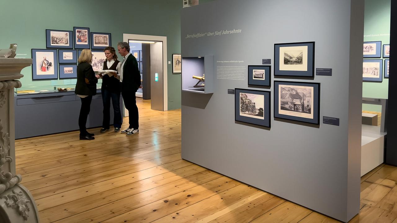 © ORF/Interspot/Philip Pürcher, Carlo Hofmann Johann-Philipp Spiegelfeld steht mit Schlossbesitzerin Angelika Meran und Schloss - & Museumsleiterin Barbara Müller in einer Ausstellung in einem modern gestalteten Museumsraum.