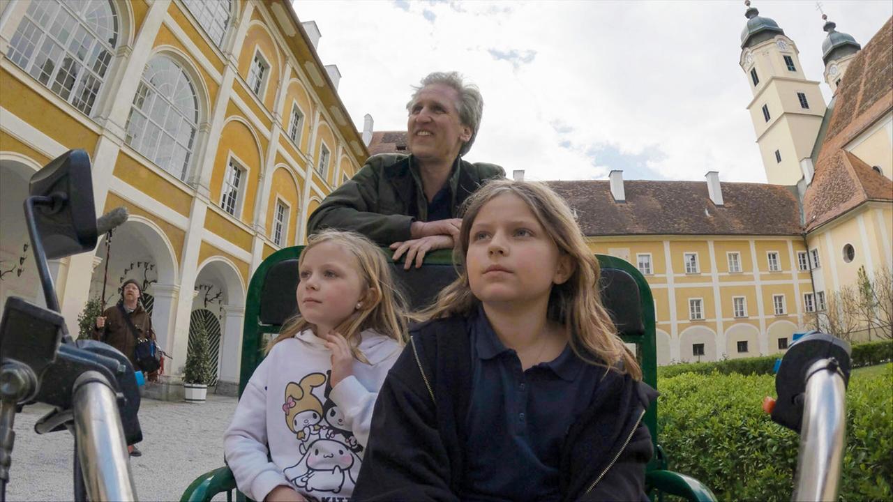 © ORF/Interspot Johann-Philipp Spiegelfeld fährt mit den zwei Kindern Franziska und Fillipa Meran in einem kleinen Gefährt durch den barocken Innenhof eines Stiftes.