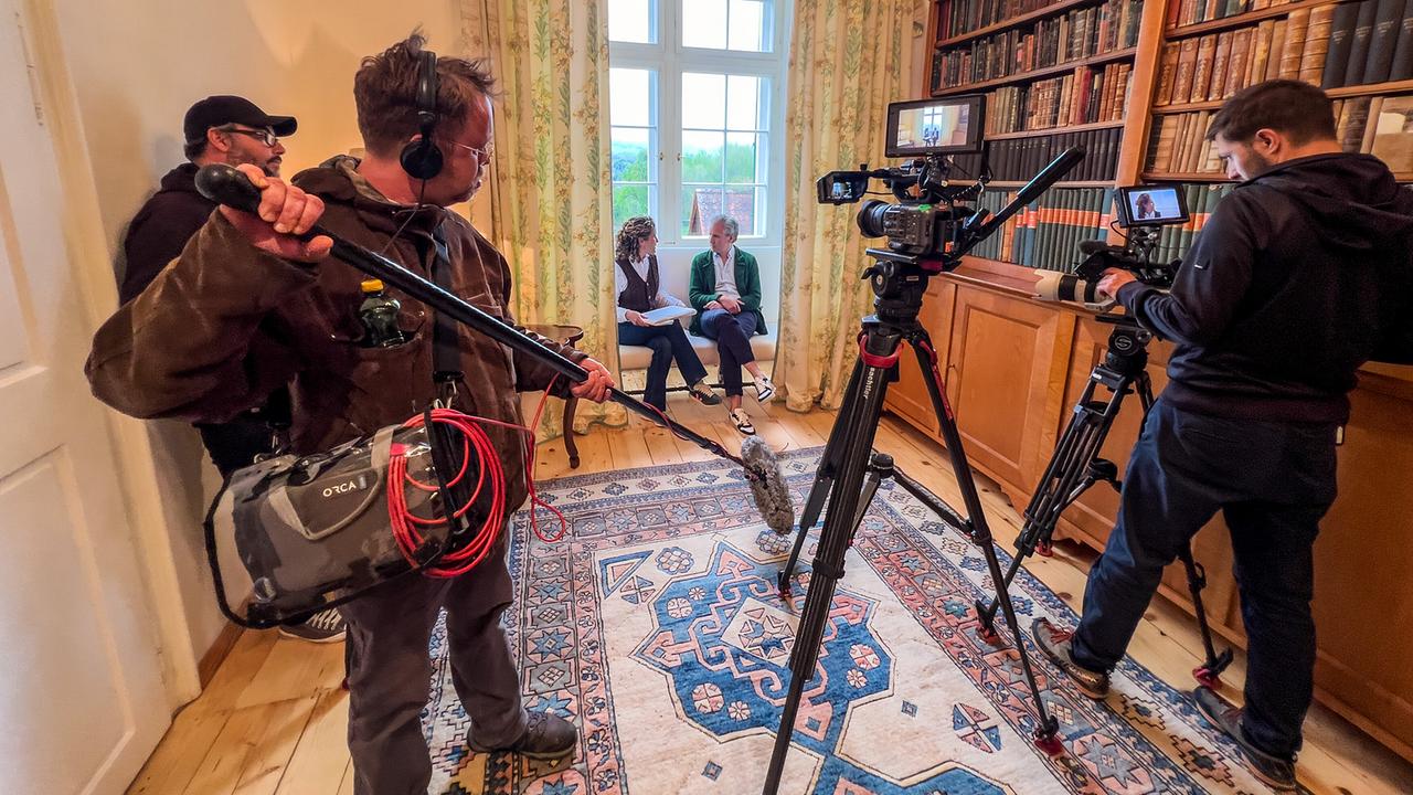 © ORF/Interspot/Philip Pürcher, Carlo Hofmann Johann-Philipp Spiegelfeld sitzt mit Schlossbesitzerin Angelika Meran auf einer Fensterbank in einer Bibliothek. Vor ihnen steht das Filmteam, das das Gespräch filmt.