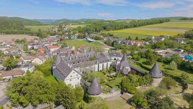 © ORF/Interspot Eine Luftaufnahme zeigt Schloss Kobersdorf mit Rundtürmen inmitten eines Dorfs und grüner Hügellandschaft.