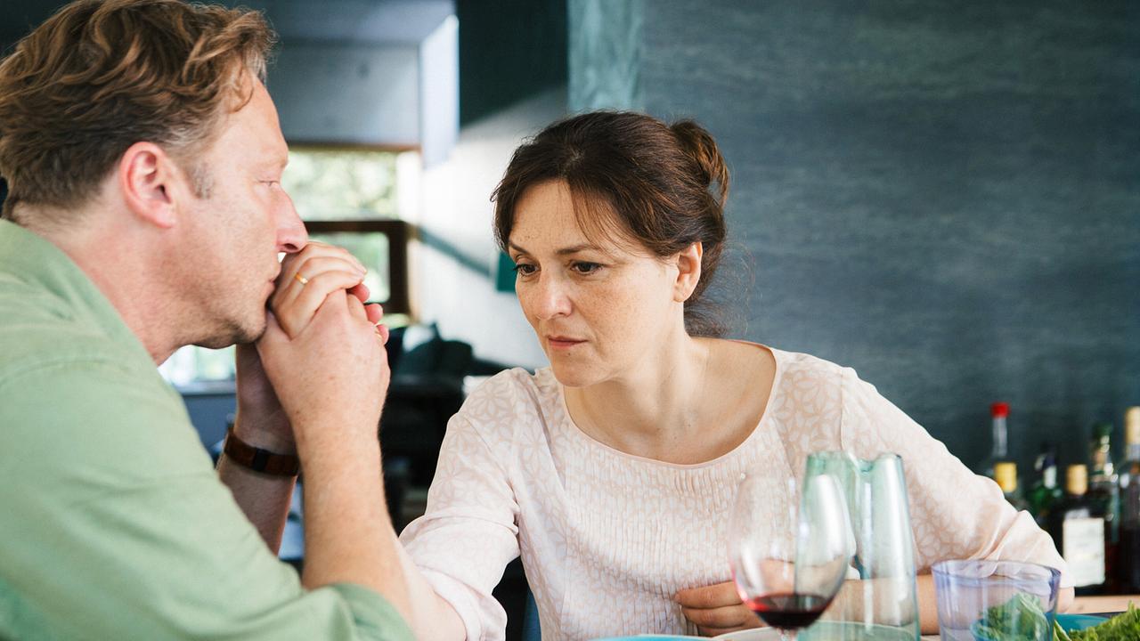 Sebastian Binder und Caroline Binder sitzen nachdenklich beim Essen am Tisch, auf dem unter anderem Wein, Gläser und Schüsseln stehen.
