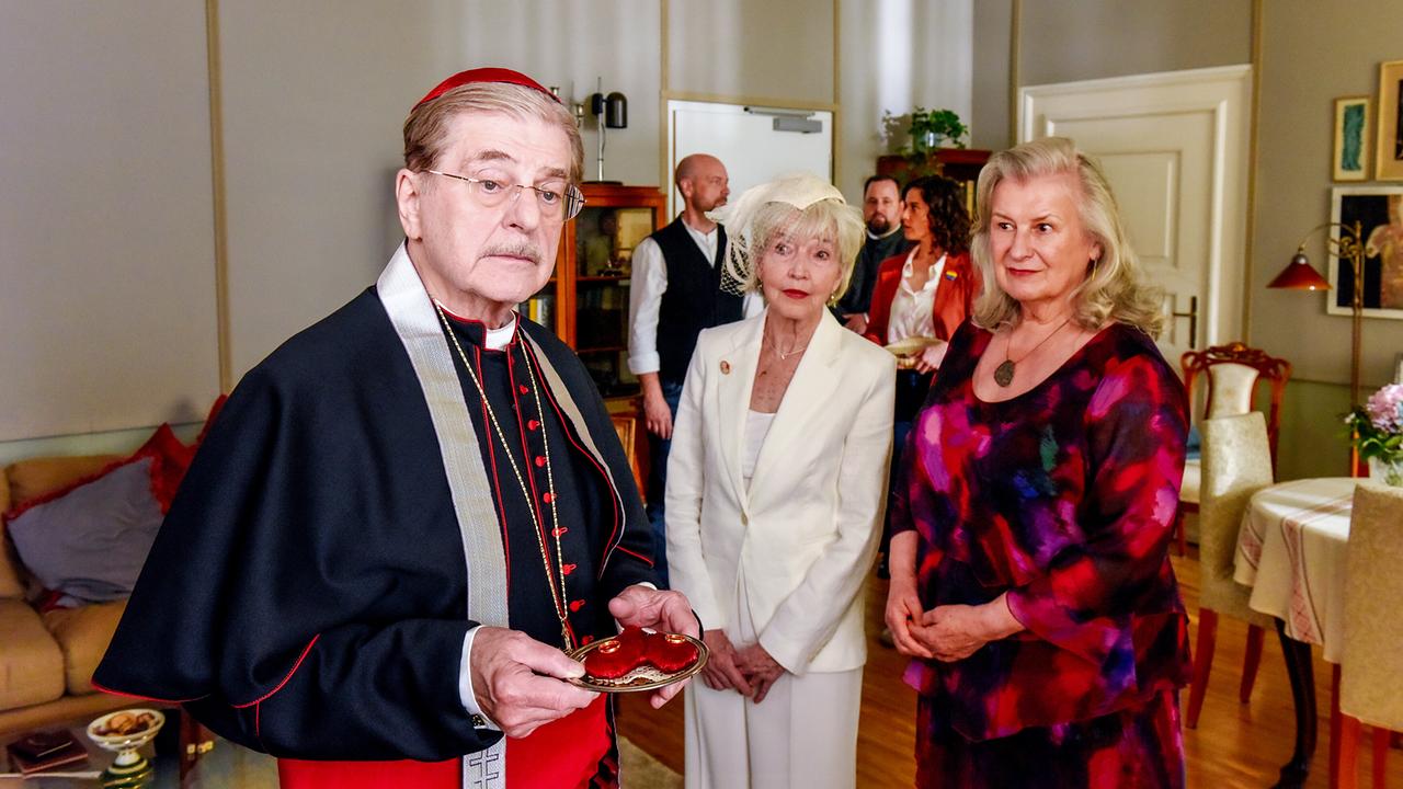 © ORF/BR/maze pictures GmbH/Barbara Bauriedl Kardinal Brunnenmayr hält in einer Wohnung ein Samttablett mit zwei Ringen, während die zwei ältere Frauen Irmi Brunnenmayr und Gertrud Hartberger neben ihm stehen. Im Hintergrund stehen Hans Reiser, Sury und Lisa Kirchberger.