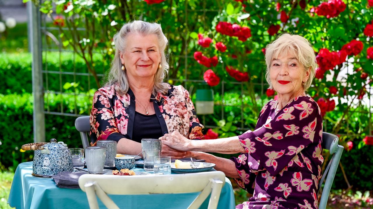 © ORF/BR/maze pictures GmbH/Barbara Bauriedl Gertrud Hartberger und Irmi Brunnenmayr sitzen in einem blühenden Rosengarten an einem gedeckten Tisch, halten einander an den Händen und lächeln.
