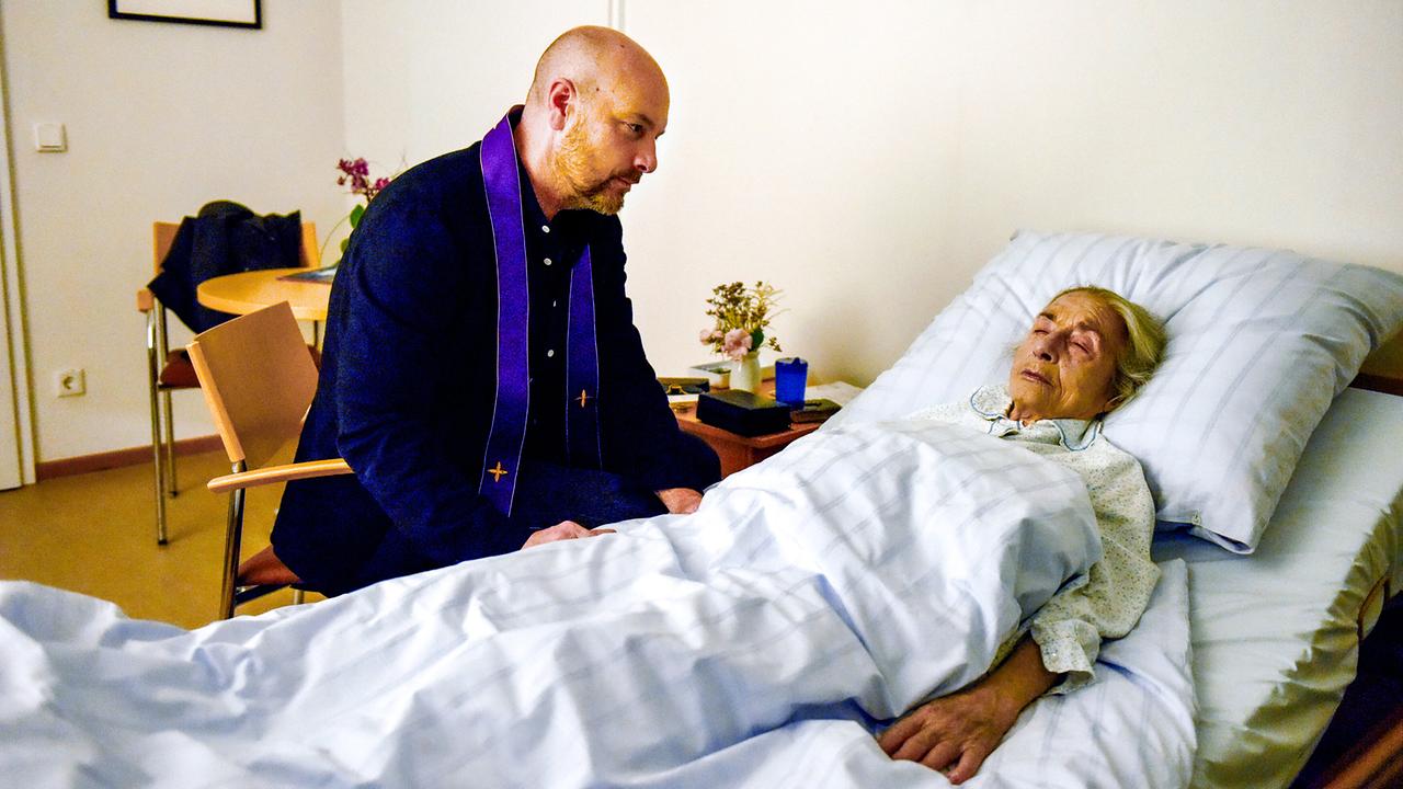 © ORF/BR/maze pictures GmbH/Barbara Bauriedl Hans Reiser mit violetter Stola sitzt am Krankenbett von Hannelore Richter und wacht still bei der älteren Frau.