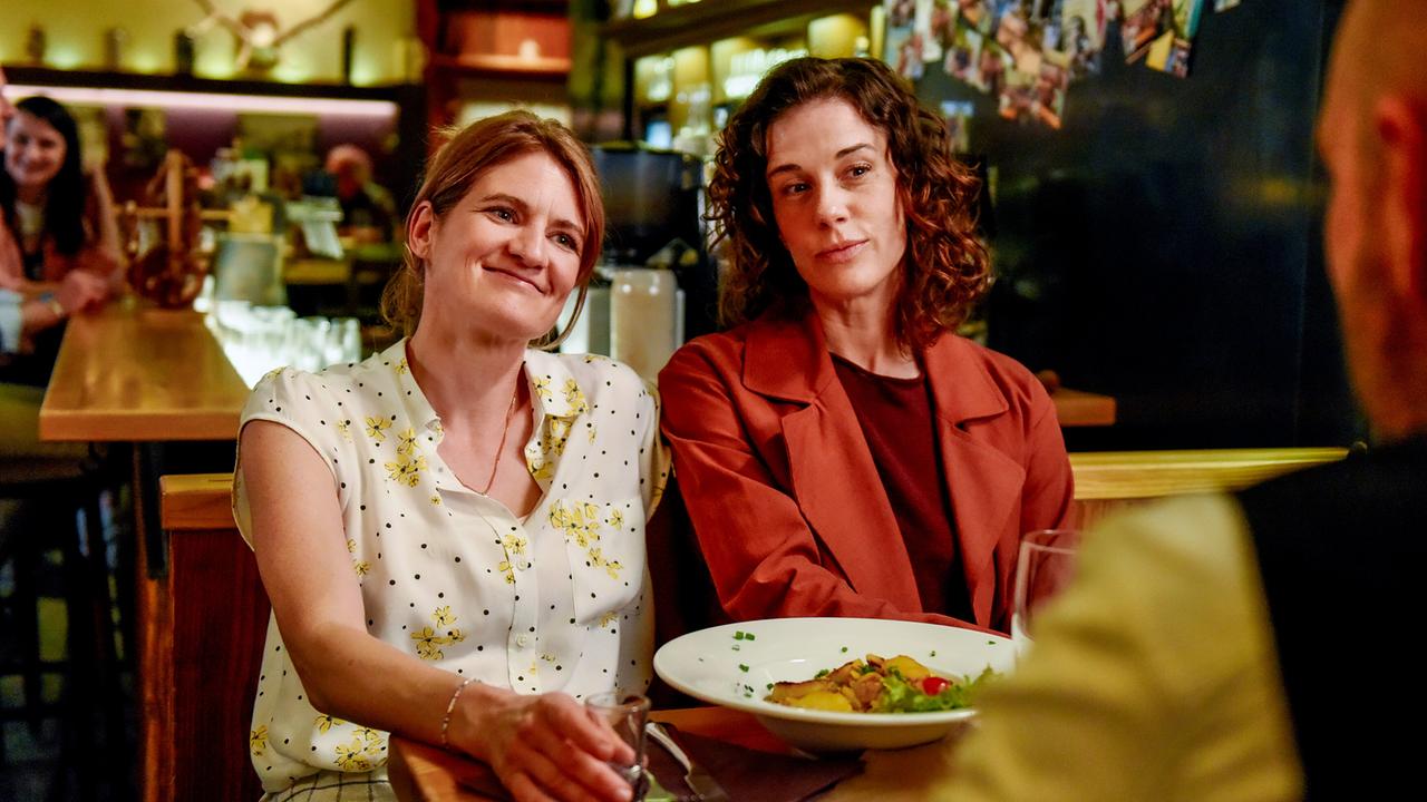 © ORF/BR/maze pictures GmbH/Barbara Bauriedl Heike Eder und Lisa Kirchberger sitzen in einem Lokal nebeneinander, schauen freundlich zu Hans Reiser, der ihnen gegenüber sitzt, und haben Essen und Getränke vor sich auf dem Tisch.