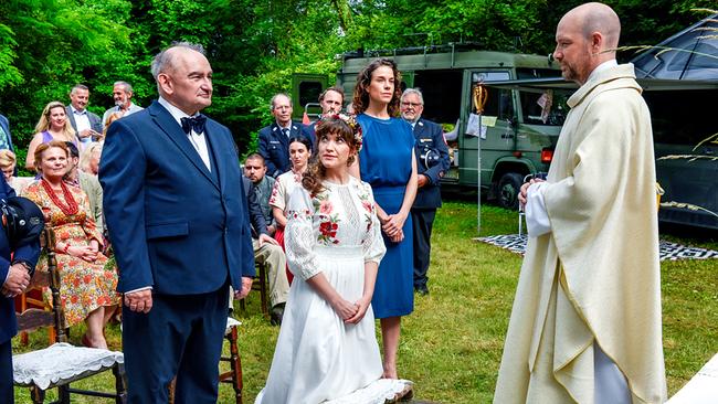 © ORF/BR/maze pictures GmbH/Barbara Bauriedl Bei einer Hochzeit im Grünen kniet Braut Agnes neben Bräutigam Michael Reiser vor Pfarrer Hans Reiser, während Gäste auf Sesseln im Hintergrund sitzen.