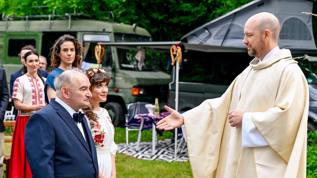 © ORF/BR/maze pictures GmbH/Barbara Bauriedl Pfarrer Hans Reiser reicht bei der Trauung im Freien dem vor ihm knienden Brautpaar Michael Reiser und Agnesa Owiecka die Hand vor Gästen und Wohnmobilen am Rand.