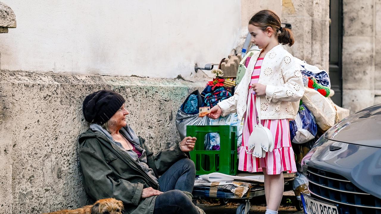 Georgina Mühlbauer, ein kleines Mädchen, gibt der obdachlosen Frau Vreni Prilmeyer auf der Straße Geld, während ein Hund daneben liegt.