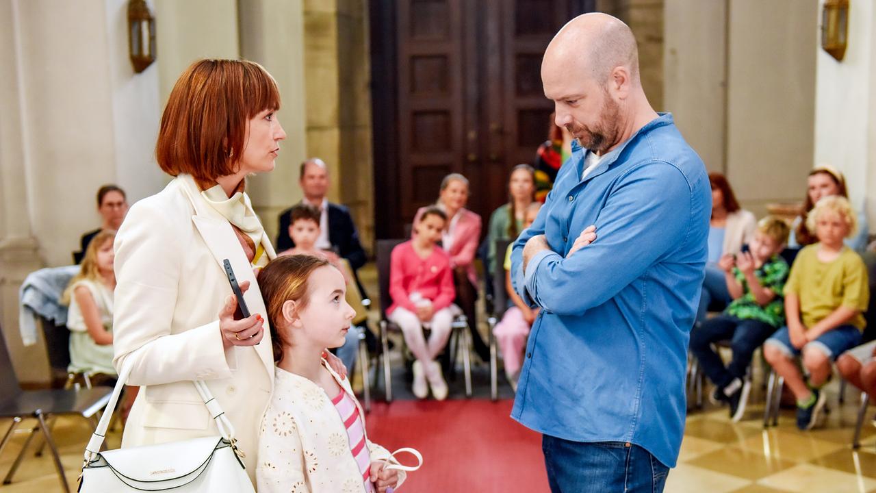 In einem Gemeindesaal stehen sich Heike Mühlbauer mit der kleinen Georgina und Hans Reiser in blauem Hemd bei einem Erstkommunionstreffen angespannt gegenüber.