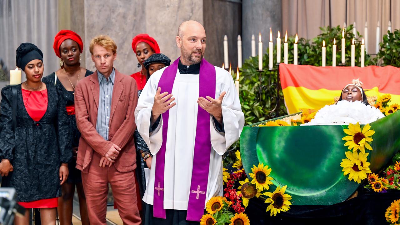 Pfarrer Reiser spricht bei einer Trauerfeier vor einem offenen, mit Blumen geschmückten Sarg zu den Anwesenden. Im Hintergrund neben dem Sarg stehen Heiner Kurz, Albina und weitere Trauergäste.