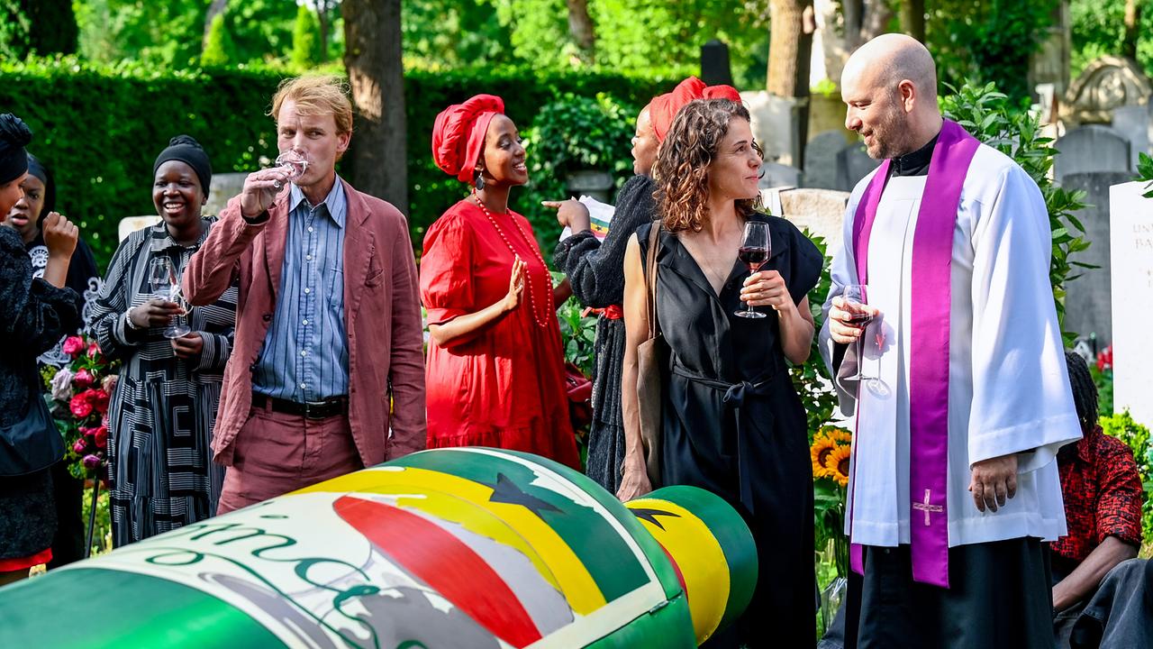 Auf einem Friedhof unterhalten sich Lisa Kirchberger,  Pfarrer Lisa Kirchberger und weitere Trauergäste mit Weingläsern vor einem bunt bemalten Sarg.