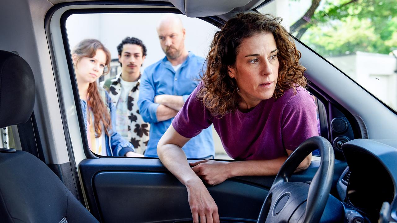 Lisa Kirchberger beugt sich durch die offene Fensterscheibe in ein Auto hinein, während Nathalie Kirchberger, Roger und Hans Reiser draußen gespannt zusehen.