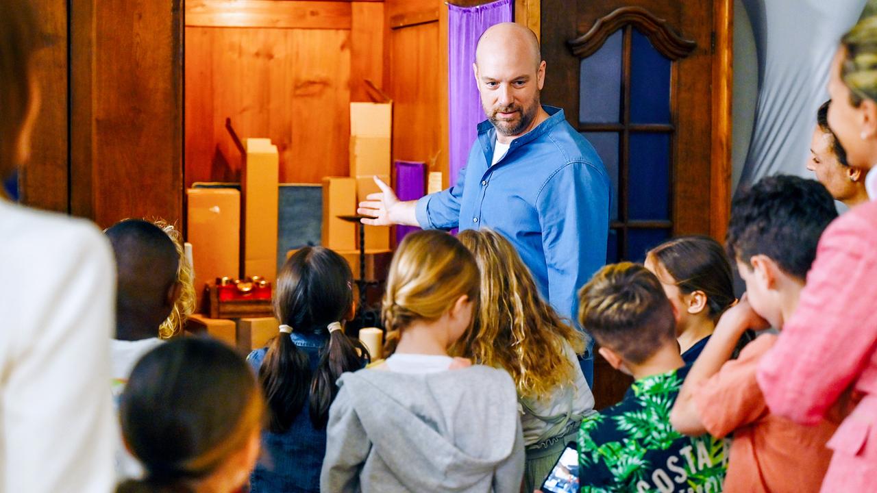 Hans Reiser erklärt einer Kindergruppe in einer Kirche etwas und zeigt dabei auf den offenen Beichtstuhl gefüllt mit Paketen im Hintergrund.