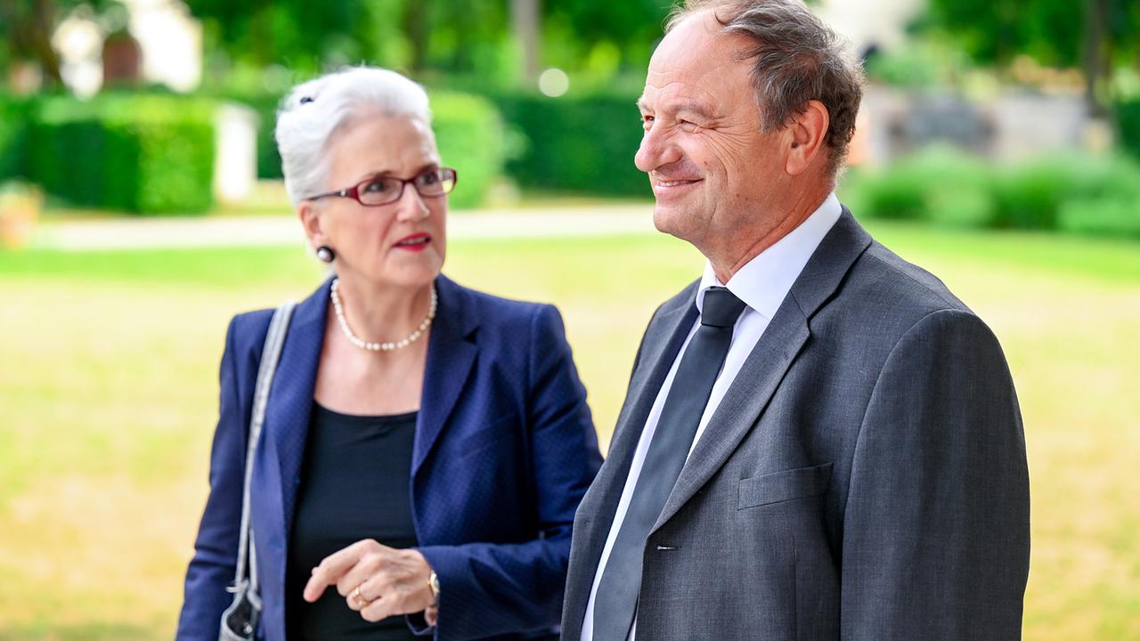 Eine elegant gekleidete Dauerwitwe und Pfarrer Schießler im Anzug stehen nebeneinander im Freien.