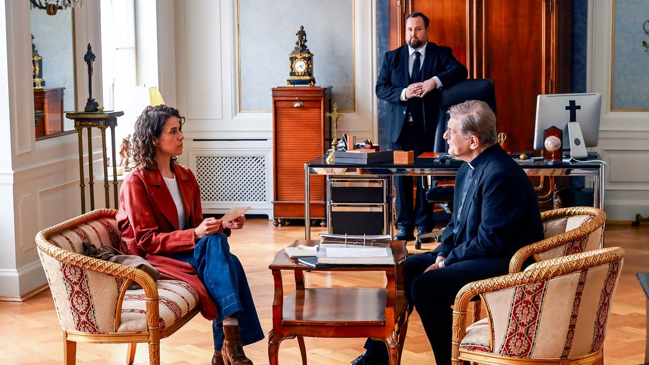 Lisa Kirchberger sitzt Kardinal Brunnenmayer in dessen Büro zu einem Gespräch gegenüber, im Hintergrund leht Sury an einem Schreibtischsessel.