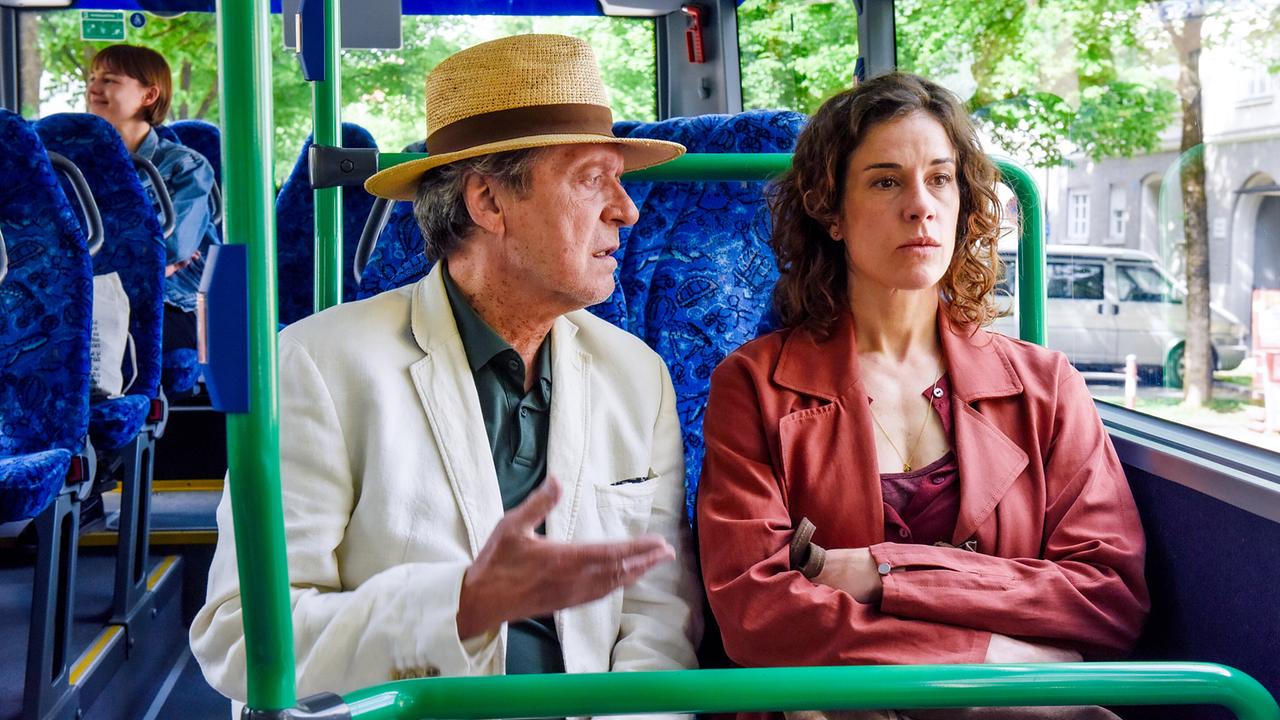 Luigi Vogelsang, ein älterer Mann im Anzug und mit Strohhut, redet im Bus mit der neben ihm sitzenden Lisa Kirchberger.