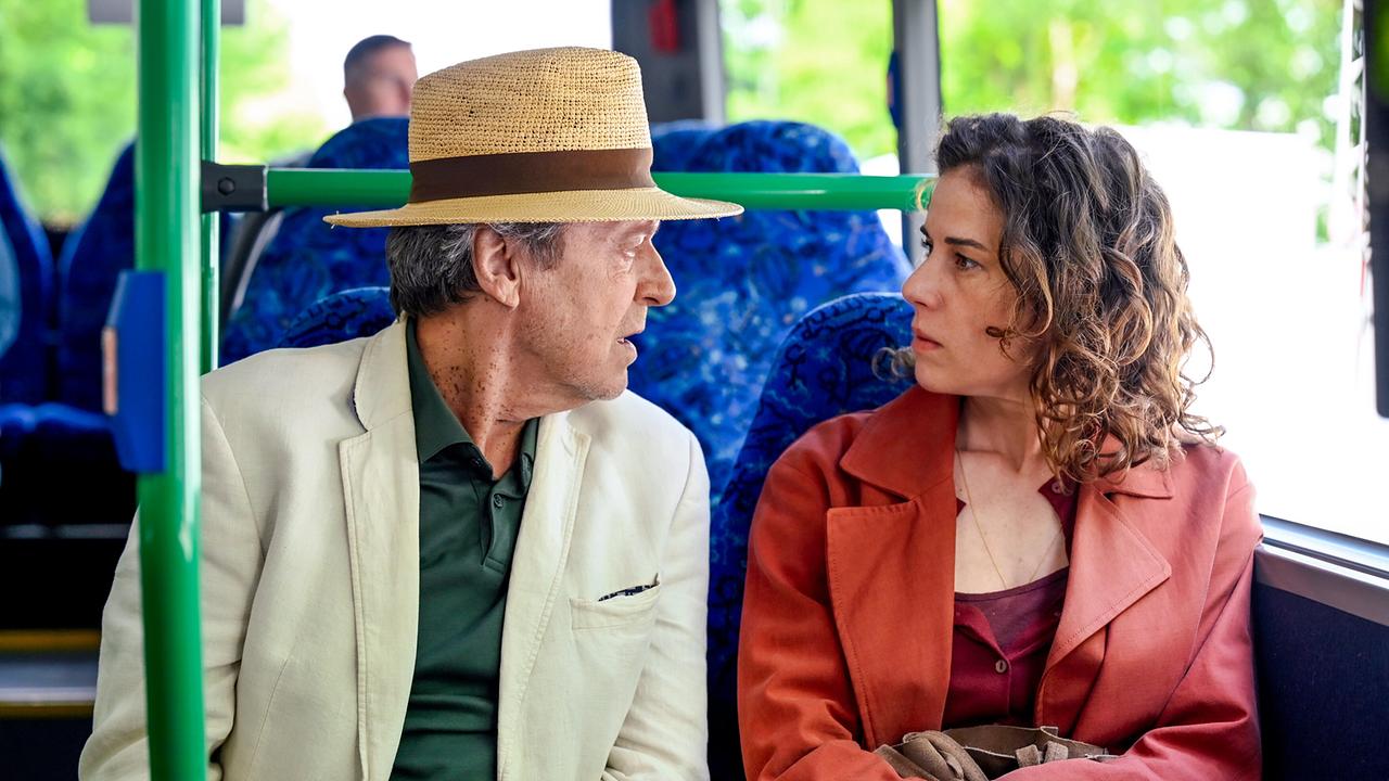 Luigi Vogelsang, ein älterer Mann im Anzug und mit Strohhut, redet im Bus mit der neben ihm sitzenden Lisa Kirchberger.