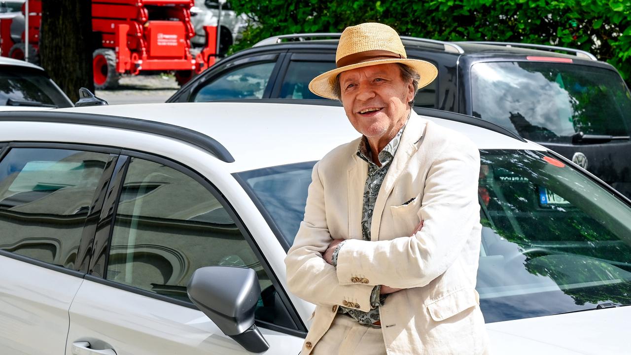 Luigi Vogelsang, ein älterer Mann im Anzug und mit Strohhut,  lehnt lächelnd mit verschränkten Armen an einem parkenden weißen Auto.