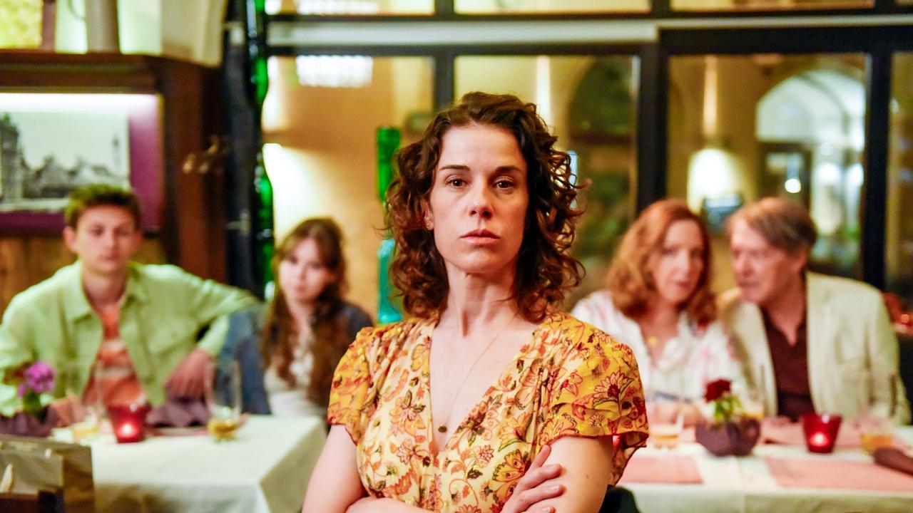 Lisa Kirchberger steht mit verschränkten Armen in einem Restaurant, während Florian und Nathalie Kirchberger und Cornelia und Luigi Vogelsang unscharf im Hintergrund an Tischen sitzen.