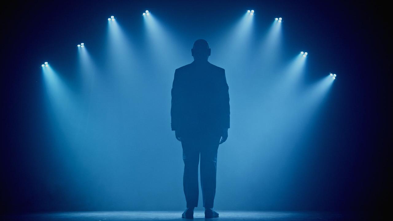 © ORF/BFILM Die Silhouette eines Mannes steht allein auf einer nebelverhangenen Bühne im Licht zahlreicher blauer Scheinwerfer.
