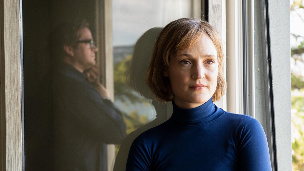 Vicky Krieps als Ingeborg Bachmann steht in einem Raum am Fenster, während sich die Silhouette von Ronald Zehrfeld (Max Frisch) im Glas spiegelt.