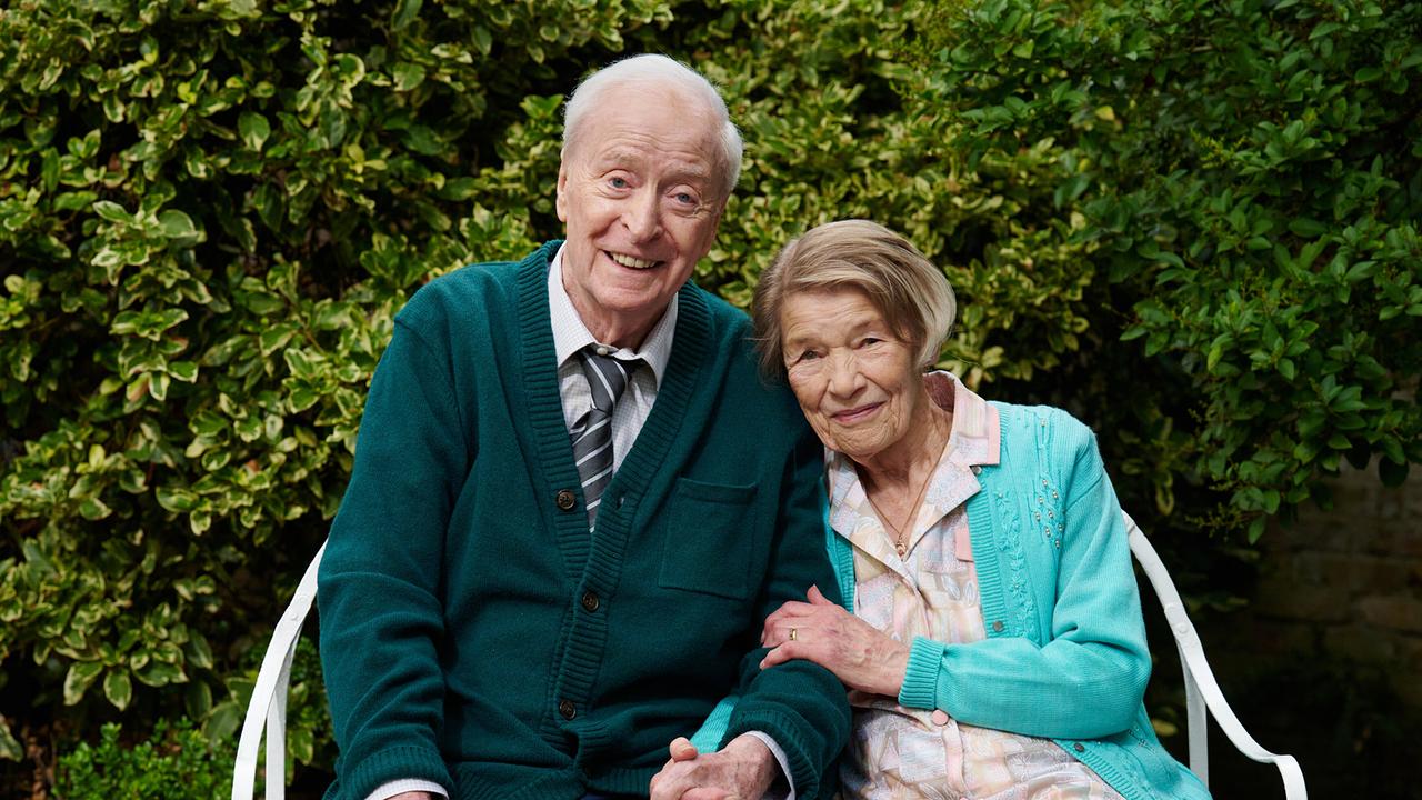 Michael Caine als Bernard Jordan und Glenda Jackson als Irene Jordan sitzt lächelnd eng beieinander auf einer Gartenbank vor einer dichten grünen Hecke.