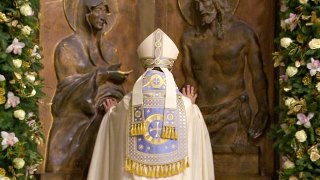 Ein Geistlicher in festlicher liturgischer Kleidung steht betend vor einem kunstvoll verzierten Relief mit religiösen Figuren, eingerahmt von üppigem Blumenschmuck.