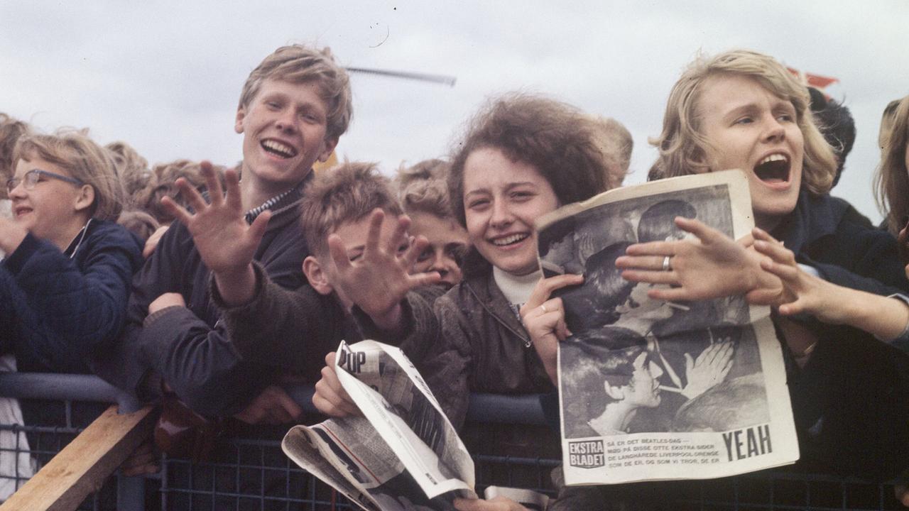 Eine dicht gedrängte Gruppe von Menschen steht hinter einer Absperrung und hält Zeitungen und Magazine in die Höhe im Rahmen der Beatles-Tournee 1960-1970
