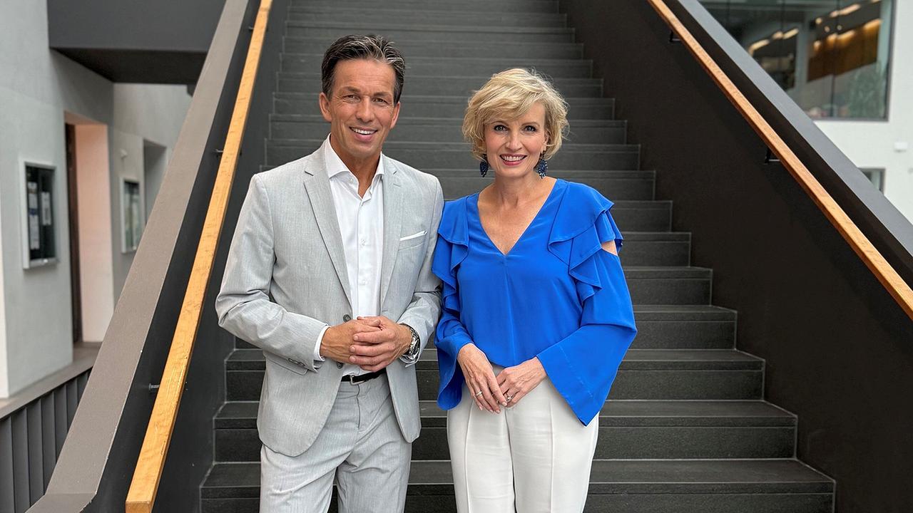 © ORF/Landesstudio Salzburg/Andrea lehner Martin Ferdiny im hellgrauen Anzug und Romy Seidl-Laux in blauer Bluse stehen lächelnd vor einer breiten Treppe in einem modernen Gebäude.